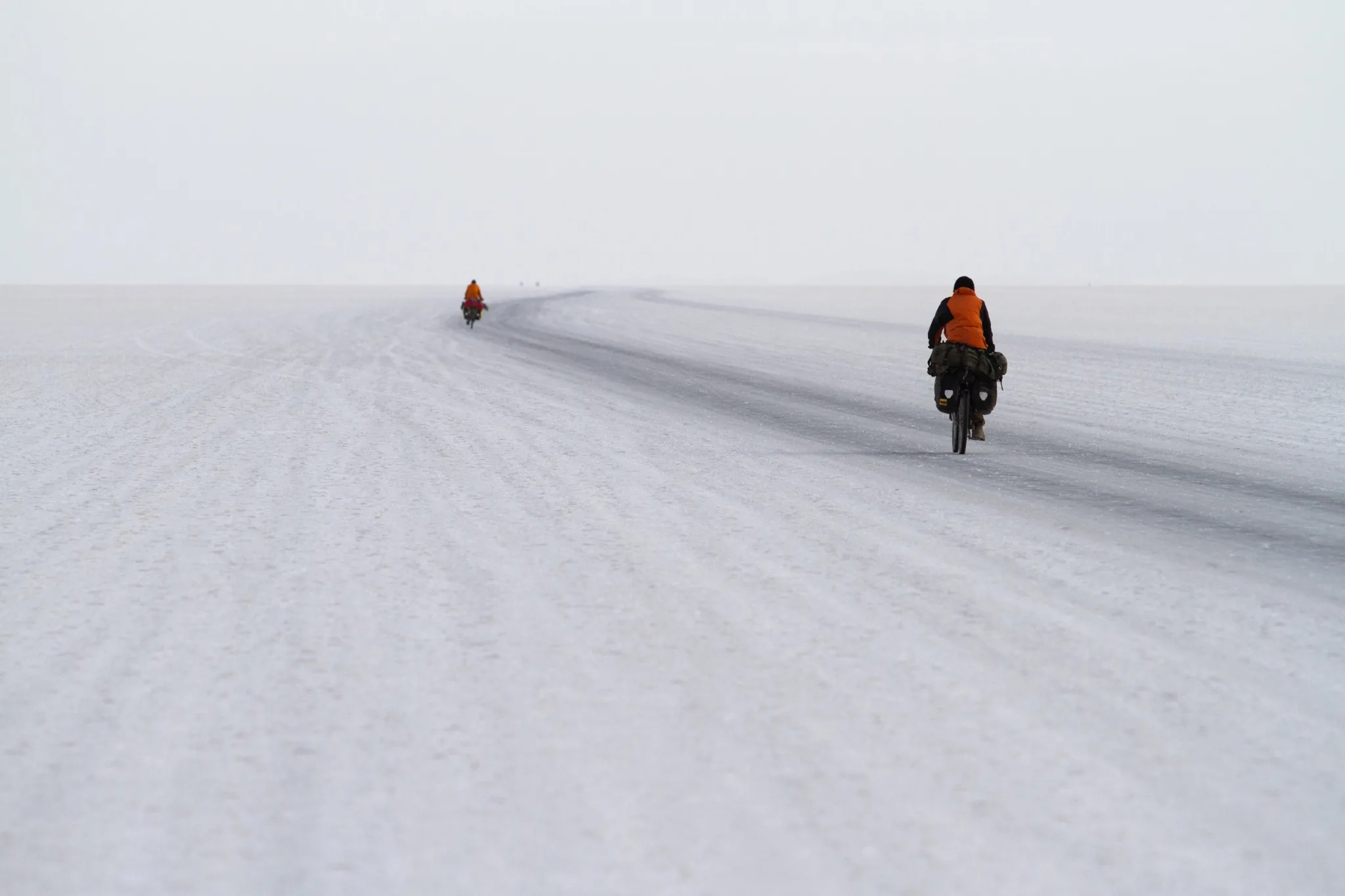 Two loaded touring cyclists crossing a broad salt flat, one riding in the foreground and another in the distance.